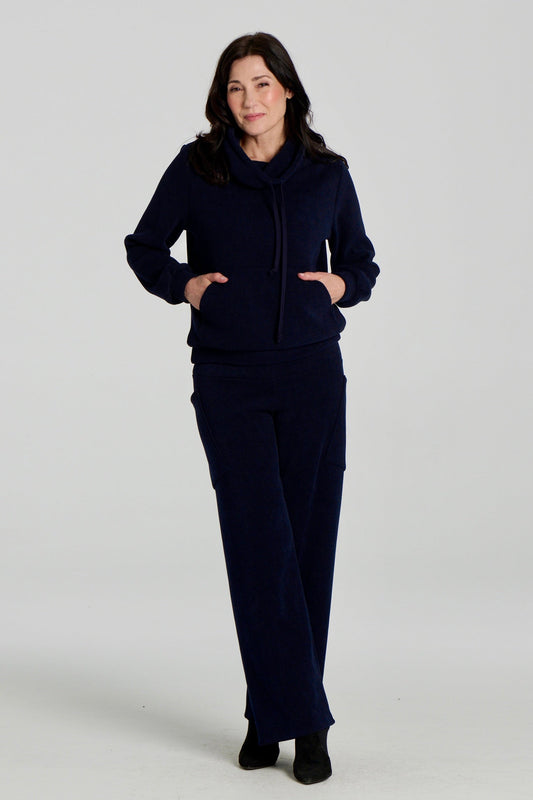 A woman wearing the Lorenzo Pants by Kollontai with the Ramos Sweater, both in a ribbed navy corduroy-like fabric. The Sweater has a large cowl neck with a drawstring and a kangaroo pocket, the pants have a wide pull on waist, straight legs, and large patch pockets. She is standing against a white background. 