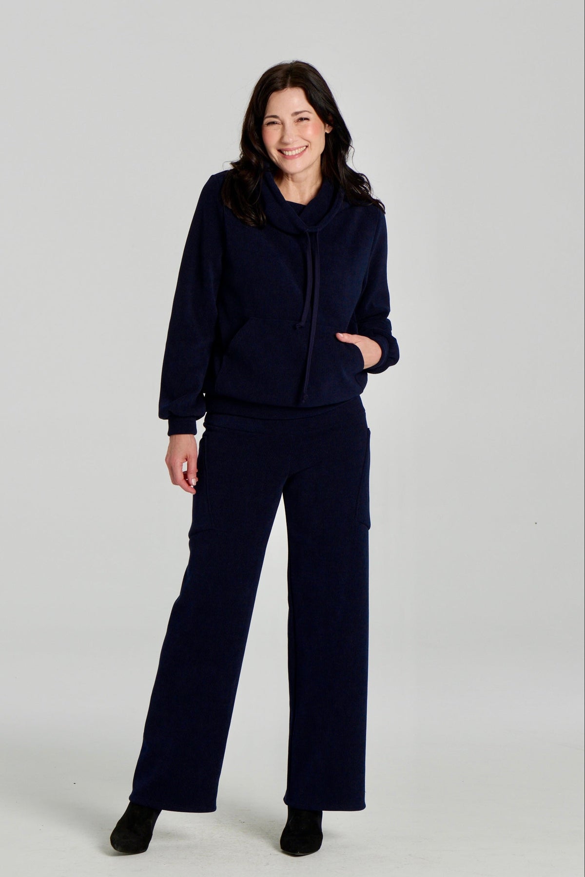 A woman wearing the Lorenzo Pants by Kollontai with the Ramos Sweater, both in a ribbed navy corduroy-like fabric. The Sweater has a large cowl neck with a drawstring and a kangaroo pocket, the pants have a wide pull on waist, straight legs, and large patch pockets. She is standing against a white background. 