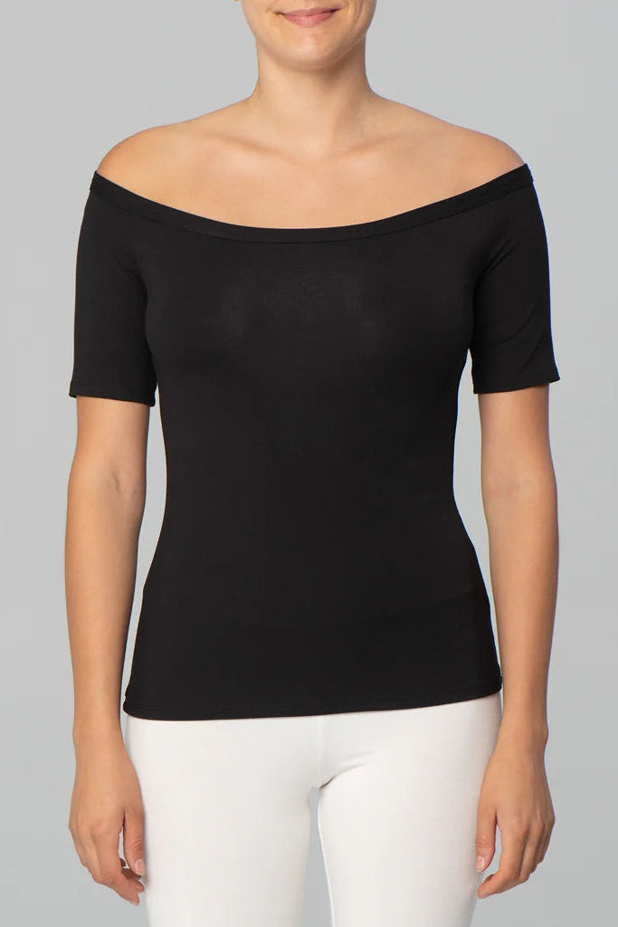 Torso view of a woman wearing the Juno Top by Kollontai in Black, an off-the-shoulder top with short sleeves. She is wearing it with white pants against a grey background.