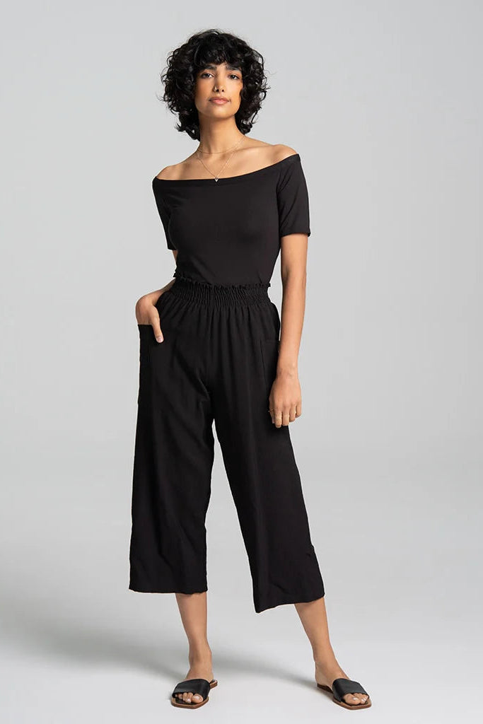 A woman wearing the Juno Top by Kollontai in Black, an off-the-shoulder top with short sleeves. She is wearing it with black wide-leg pants against a grey background.