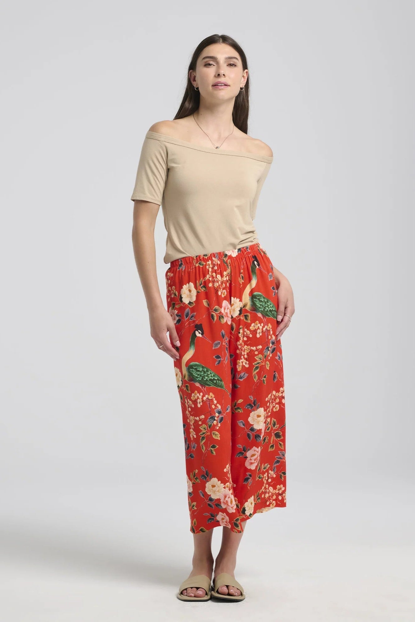 A woman wearing the Paraiso Pants by Kollontai in Red floral, with an elastic waist, wide cropped legs, and side slits at the hems. She's wearing them with a beige top and is standing in front of a white background. 