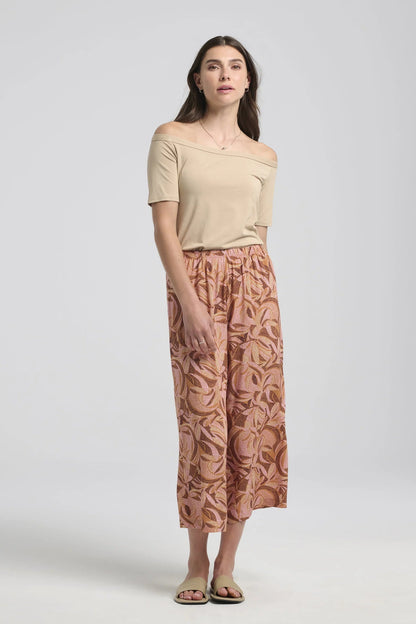 A woman wearing the Isiclora Pants by Kollontai in Cinnamon print, a lightweight pant with an elastic waist and flowing, cropped legs. She is wearing them with a beige top and standing in front of a white background. 