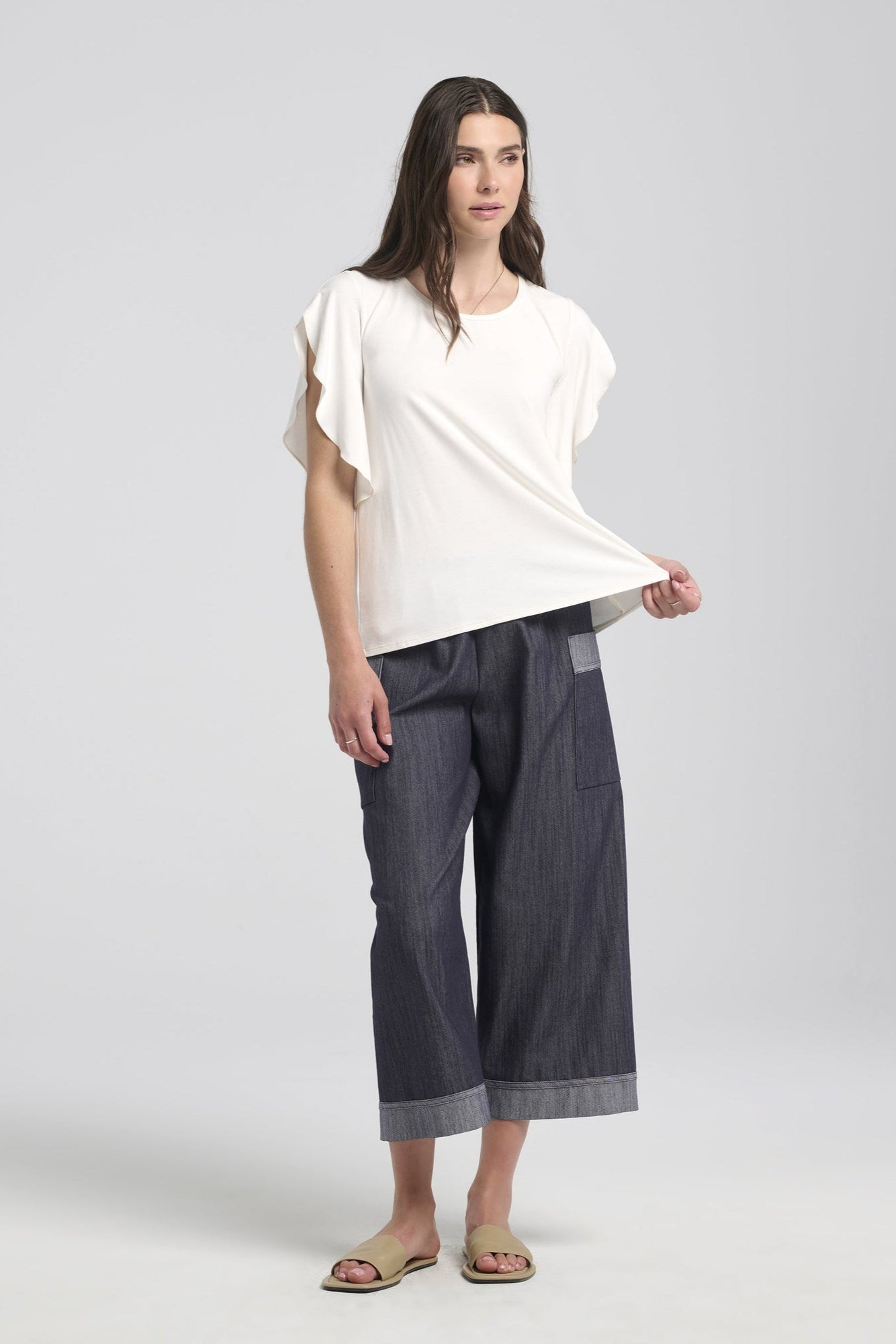 A woman wearing the Morwenna Pants from Kollontai, lightweight denim pants with wide cropped legs, an elastic waist, and contrasting trim on the large patch pockets and the cuffs. She is wearing them with the Morgana T-Shirt in white and is standing in front of a white background. 