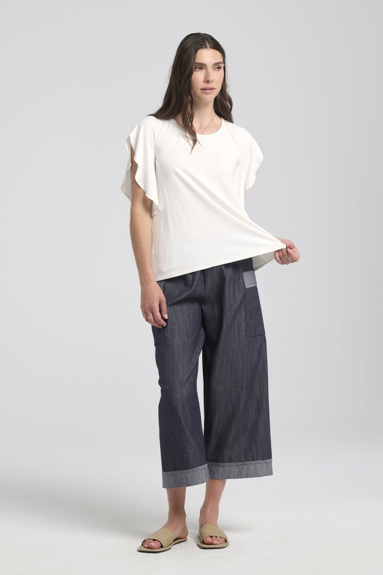 A woman wearing the Morgana T-Shirt by Kollontai in White, a loose fitting top with a round neck and fly-away sleeves that fall to the elbow. She is wearing it with the Morwenna Pants and is standing in front of a white background. 