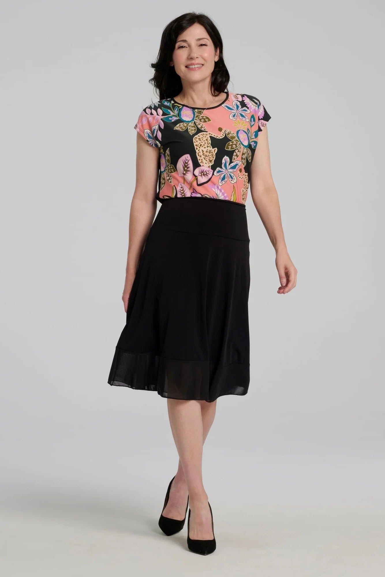 A woman wearing the Badia T-shirt by Kollontai in Bubblegum, featuring a patterned front with leopards, flowers and birds and a solid black back. It has a cap sleeves and a hi-low hem She i swearing it with a black skirt and standing in front of a grey background. 