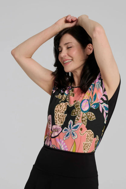 A woman wearing the Badia T-shirt by Kollontai in Bubblegum, featuring a patterned front with leopards, flowers and birds and a solid black back. It has a cap sleeves and a hi-low hem She i swearing it with black bottoms and standing in front of a grey background. 