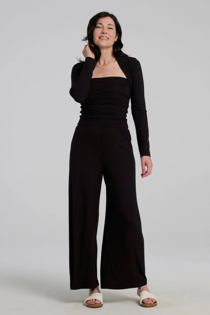 A woman wearing the Osimo Bolero by Kollontai in Black with the Dolora Jumpsuit, standing against a grey background. 