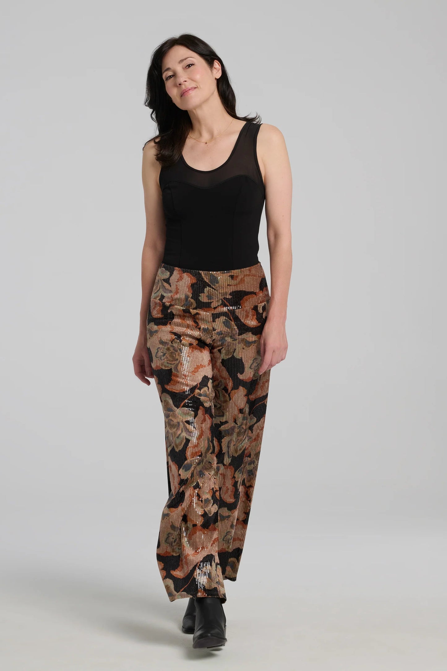 A woman wearing the Carina Pants by Kollontai, featuring floral velvet in autumn tones with clear sequins sewn right into the fabric. They have full-length straight legs and a wide pull-on waistband. She's wearing them with a black tank and standing in front of a grey background. 