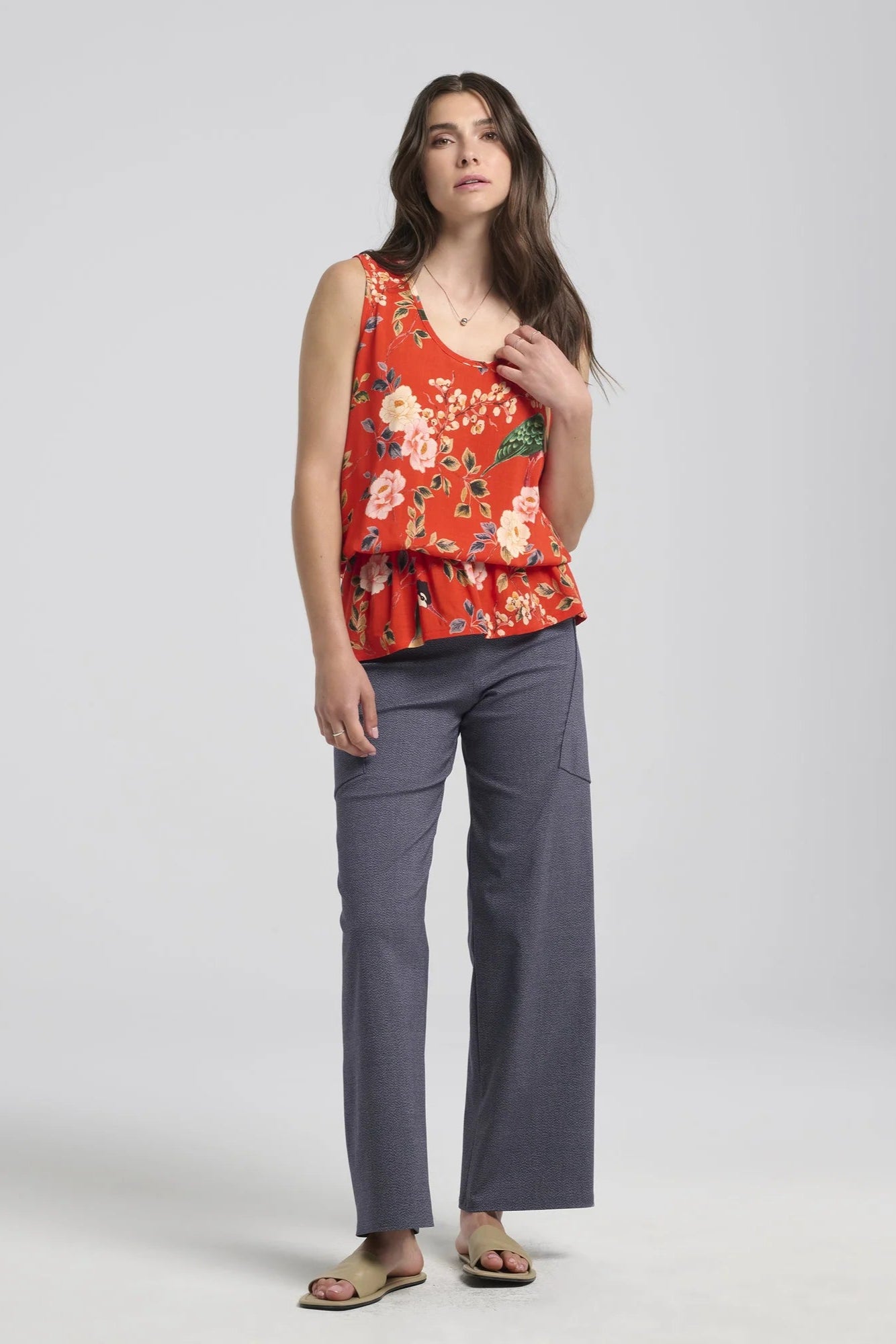 A woman wearing the Yvaine Top by Kollontai in Red, a sleeveless top with a scoop neck, wide straps, an elastic waist that creates a bloused effect. She is wearing it with the Frydek Pants in Denim and standing in front of a white background. 