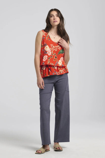 A woman wearing the Yvaine Top by Kollontai in Red, a sleeveless top with a scoop neck, wide straps, an elastic waist that creates a bloused effect. She is wearing it with the Frydek Pants in Denim and standing in front of a white background. 
