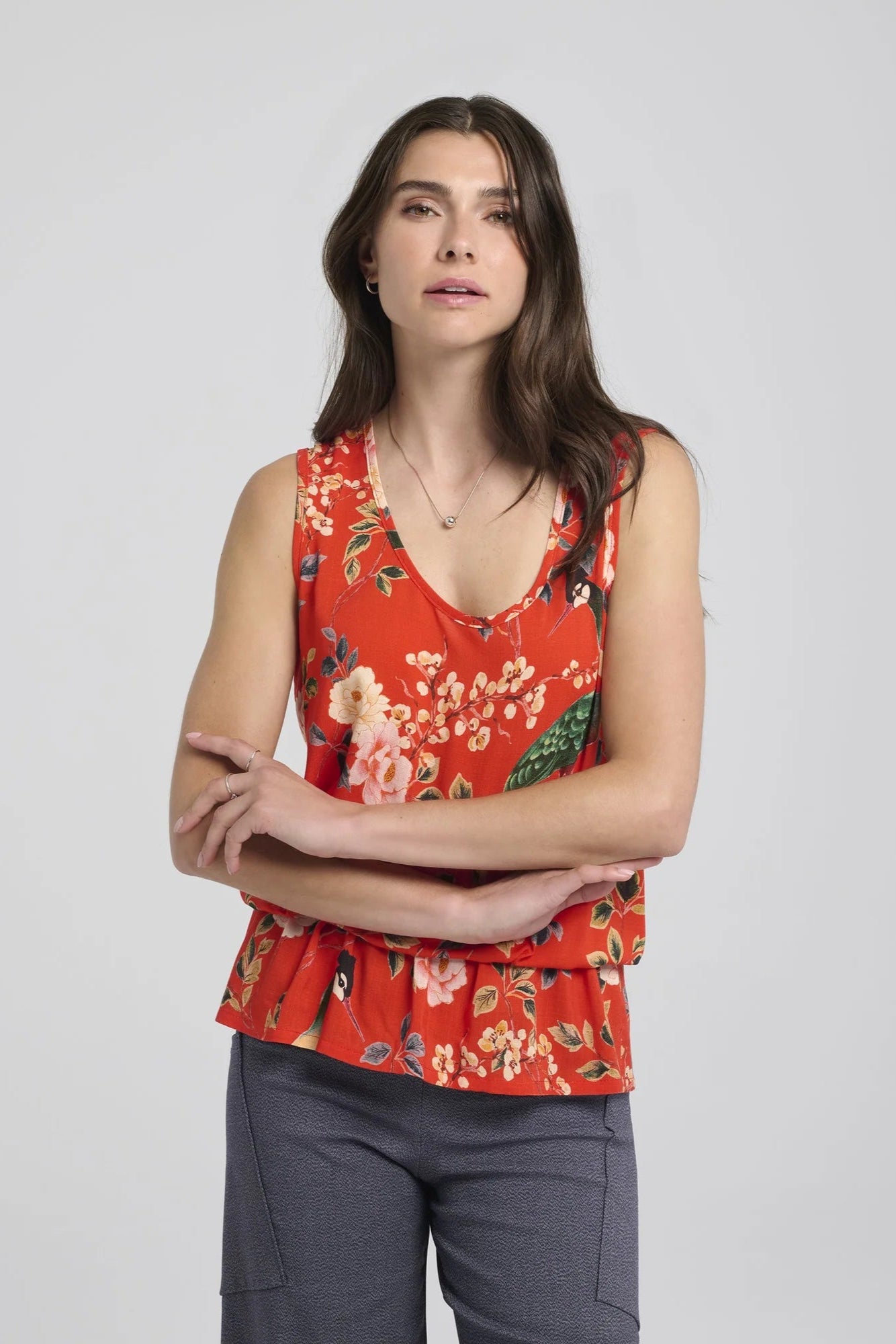3/4 shot of a woman wearing the Yvaine Top by Kollontai in Red, a sleeveless top with a scoop neck, wide straps, an elastic waist that creates a bloused effect. She is wearing it with the Frydek Pants in Denim and standing in front of a white background. 
