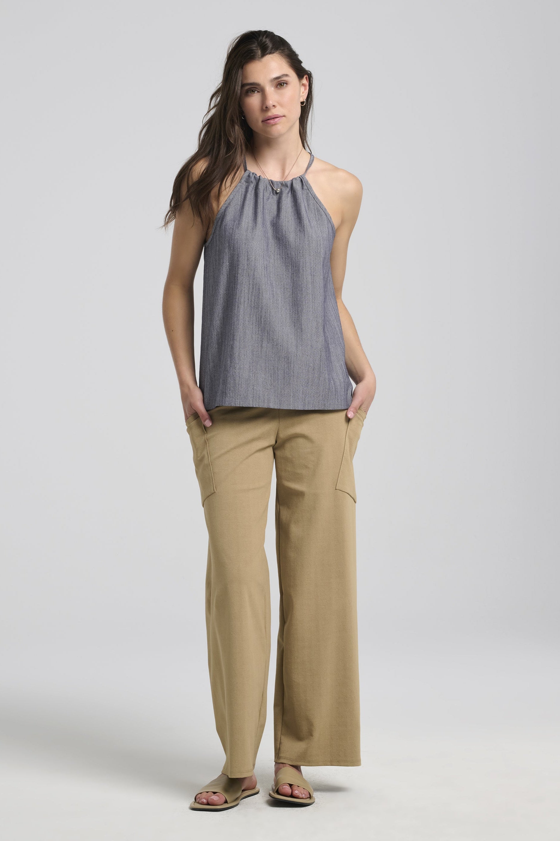 A woman wearing the Semira Camisole by Kollontai in denim, a lightweight top with an adjustable halter neck and a loose fit. She is wearing it with tan pants and is standing in front of a white background. 