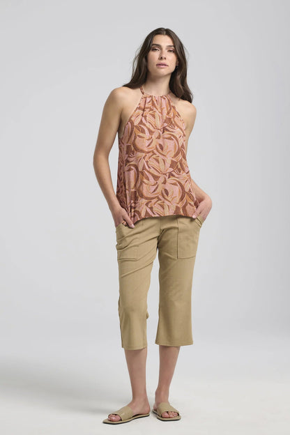 A woman wearing the Semira Camisole by Kollontai in Cinnamon print, a lightweight top with an adjustable halter neck and a loose fit. She is wearing it with tan capris and is standing in front of a white background. 