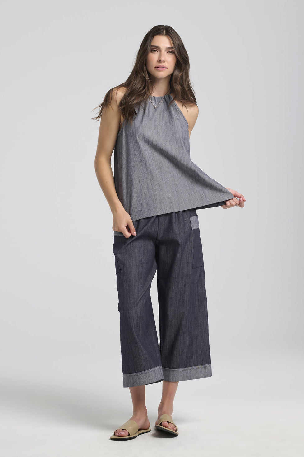 A woman wearing the Morwenna Pants from Kollontai, lightweight denim pants with wide cropped legs, an elastic waist, and contrasting trim on the large patch pockets and the cuffs. She is wearing them with the Semira Camisole and is standing in front of a white background. 