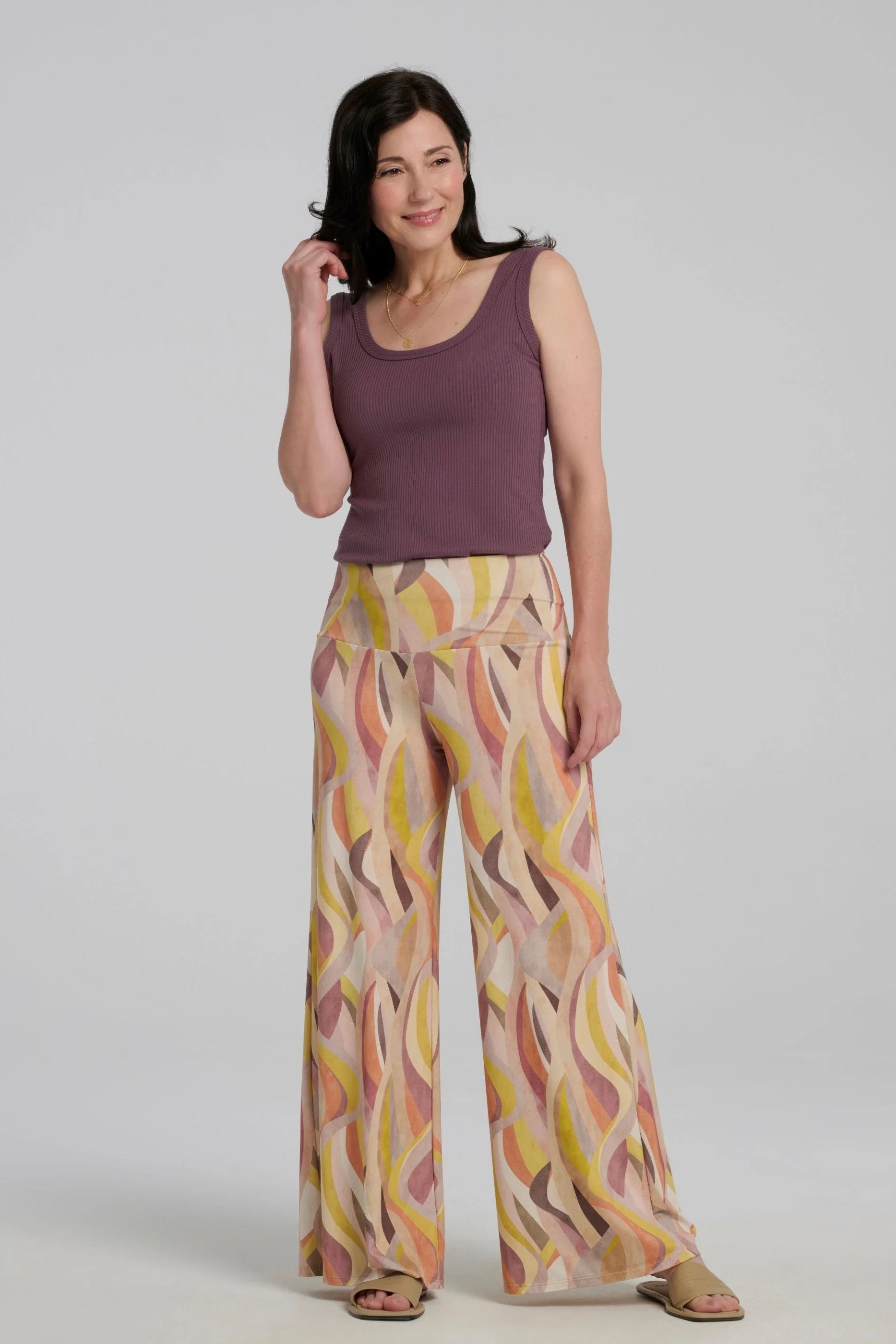 A woman wearing the Sorela pants from Kollontain in Peach print, with a wide pull-on waistband, flowing legs, and swirling geometric print. She is wearing them with a purple tank and standing in front of a white background. 