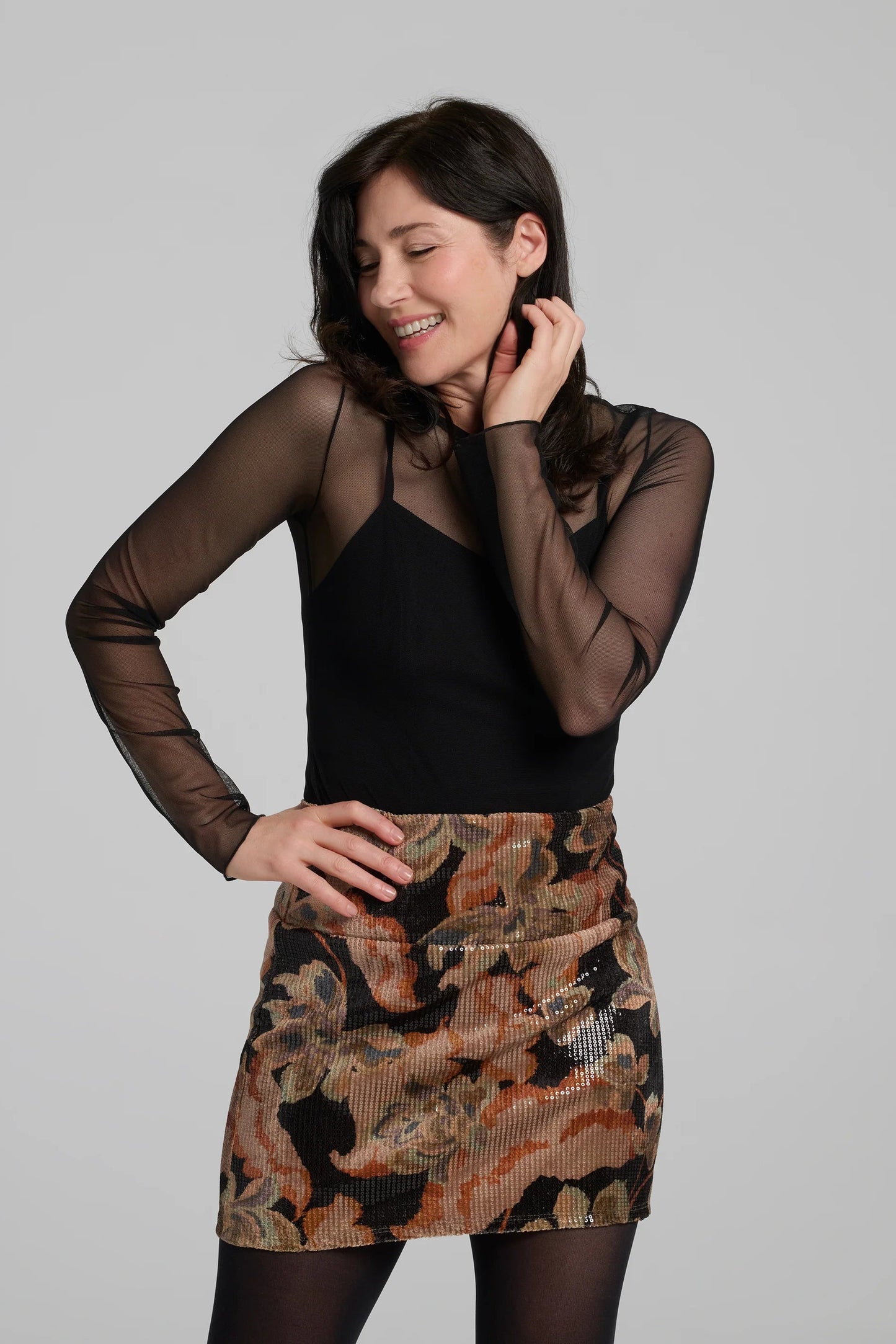 A woman wearing the Soleste Skirt by Kollontai, a floral velvet miniskirt in autumn tones with clear sequins sewn right into the fabric. It has a wide pull-on waistband. She is wearing it with a black top and standing in front of a grey background. 