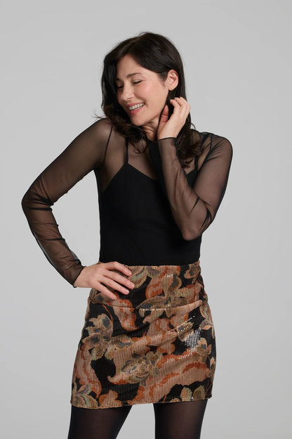 A woman wearing the Soleste Skirt by Kollontai, a floral velvet miniskirt in autumn tones with clear sequins sewn right into the fabric. It has a wide pull-on waistband. She is wearing it with a black top and standing in front of a grey background. 