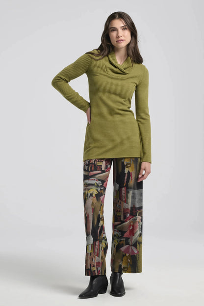 A woman wearing the Joanna Pants by Kollontai, a straight cut pant with a wide pull-on waistband and a streetscape pattern. She is wearing it with a lime green top and a white sweater, and is standing in front of a white background. 