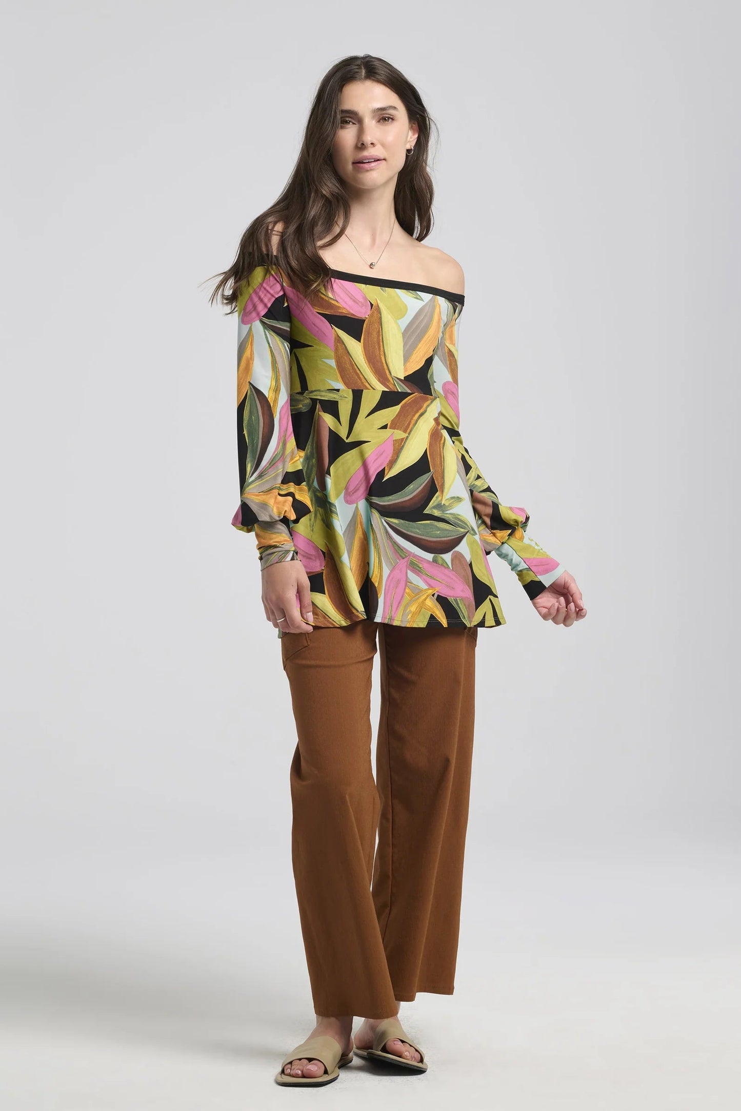 A woman wearing the Frydek Pants by Kollontai in Cinnamon, featuring wide legs, a wide pull-on waistband, and angled patch pockets. She is wearing them with an abstract print top and standing in front of a white background. 