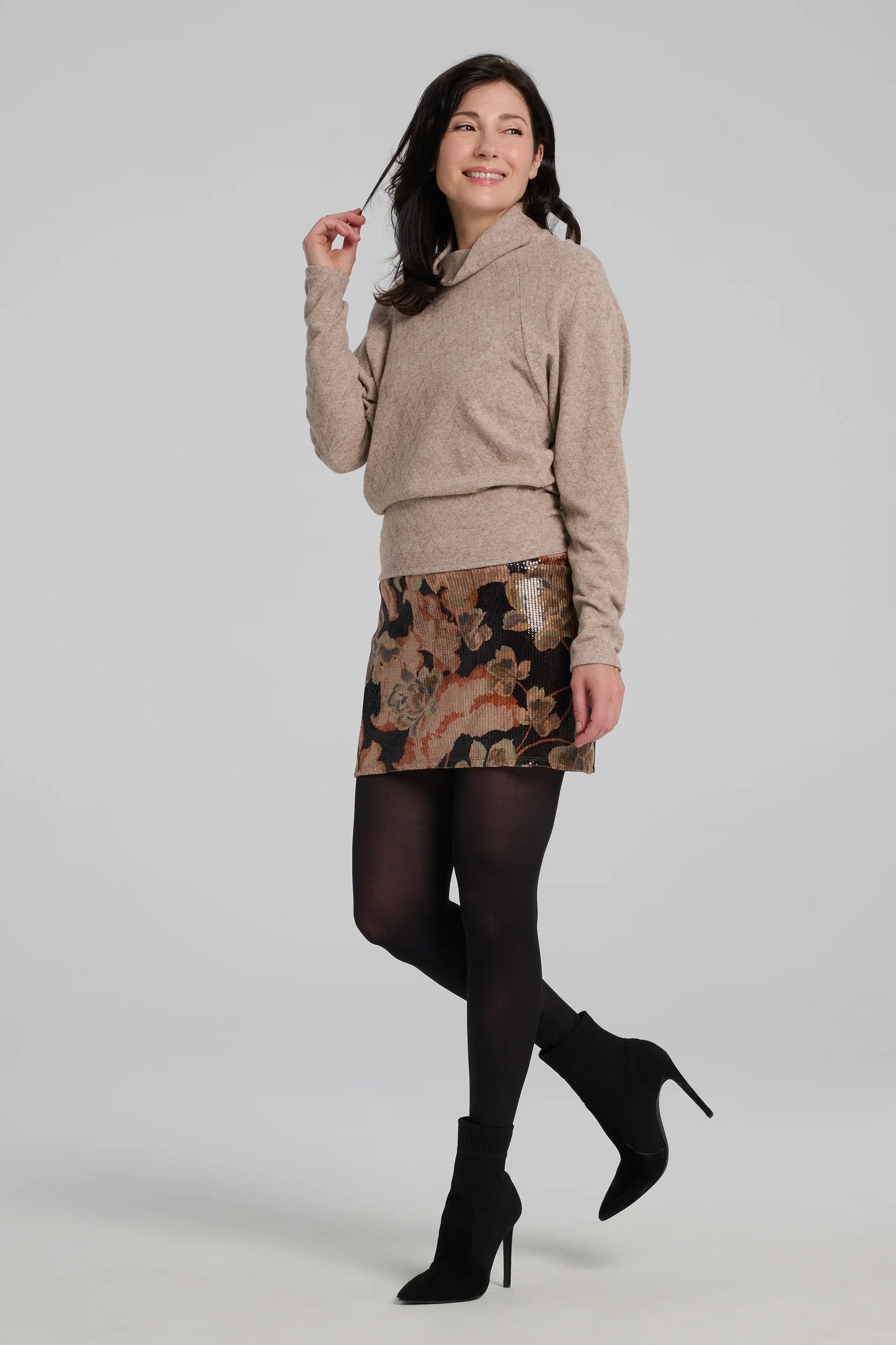 A woman wearing the Soleste Skirt by Kollontai, a floral velvet miniskirt in autumn tones with clear sequins sewn right into the fabric. It has a wide pull-on waistband. She is wearing it with a beige sweater and standing in front of a grey background. 