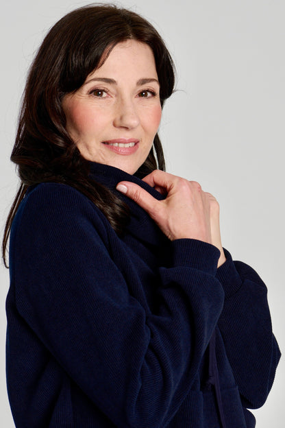 Close up side view of a woman wearing the Ramos Sweater by Kollonta in a ribbed navy corduroy-like fabric. The Sweater has a large cowl neck with a drawstring and a kangaroo pocket. She is standing against a white background.