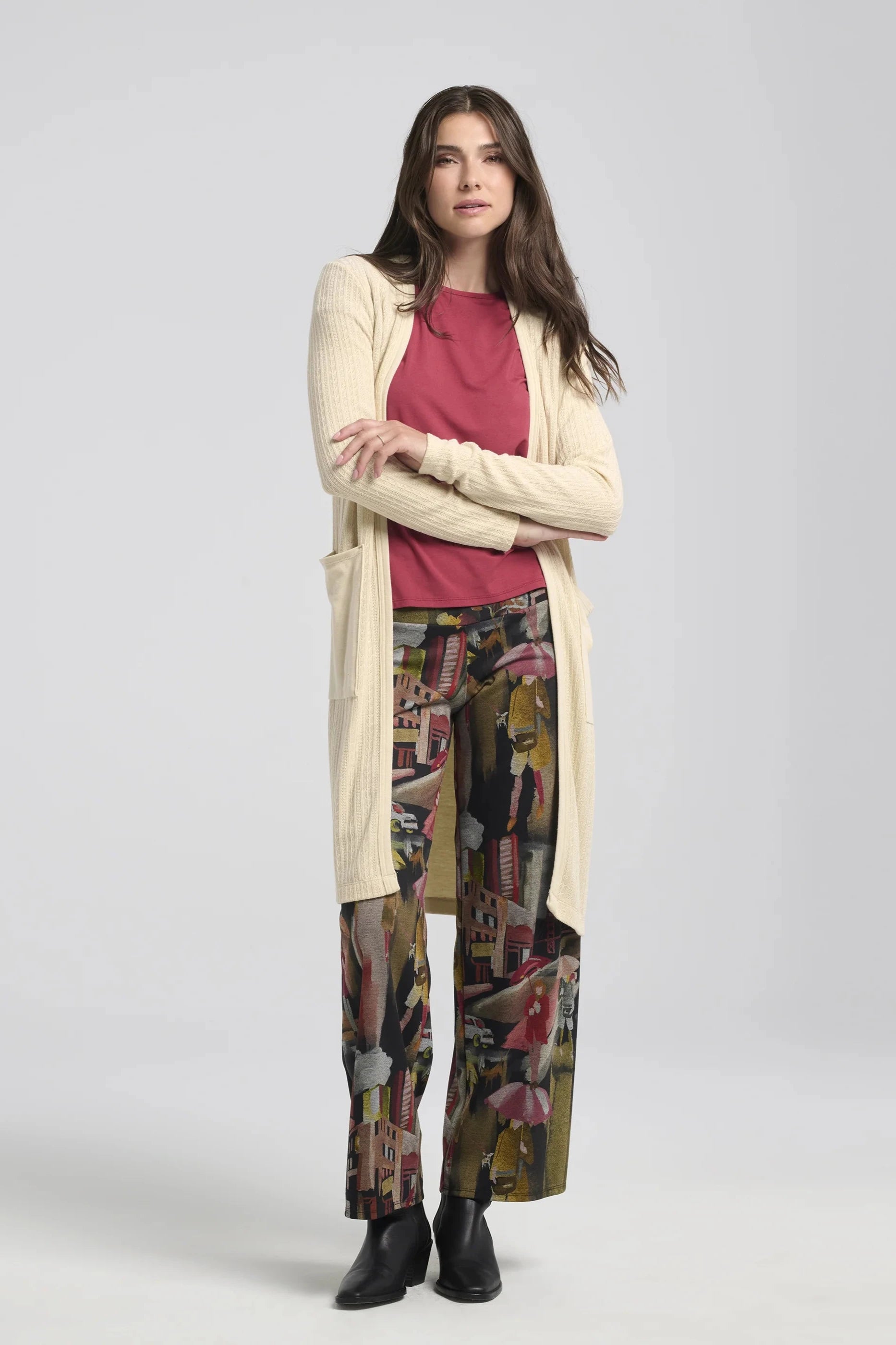 A woman wearing the Joanna Pants by Kollontai, a straight cut pant with a wide pull-on waistband and a streetscape pattern. She is wearing it with a pink top and a white sweater, and is standing in front of a white background. 