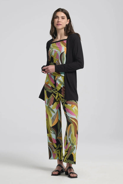 A woman wearing the Anthurium Jumpsuit by Kollontai in a Multicolour tropical print, with an off the shoulder neckline, bloused elastic waist, short sleeves and wide 7/8 length legs. She is wearing it with a black cardigan and standing in front of a grey background. 