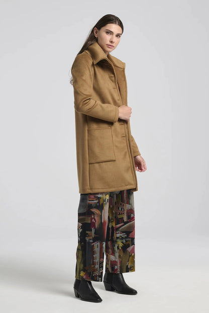 A woman wearing the Joanna Pants by Kollontai, a straight cut pant with a wide pull-on waistband and a streetscape pattern. She is wearing it with brown coat, and is standing in front of a white background. 