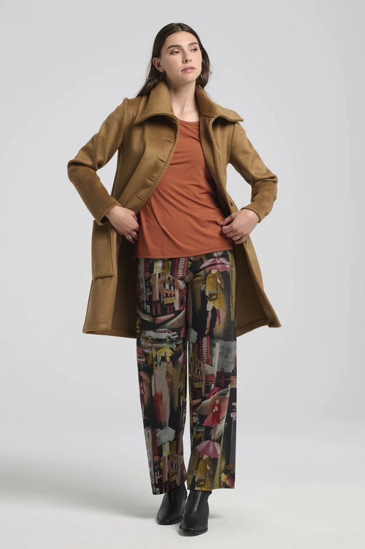 A woman wearing the Joanna Pants by Kollontai, a straight cut pant with a wide pull-on waistband and a streetscape pattern. She is wearing it with an orange top and a tan coat, and is standing in front of a white background. 