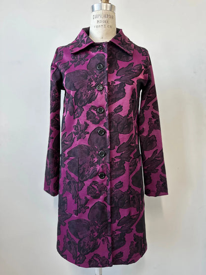 The Amarante Coat by Kollontai in Blackberry is shown on a mannequin against a white background. It's a satin coat with an intricate floral pattern, a classic collar, button front, straight cut that hits above the knee, and large pockets. 