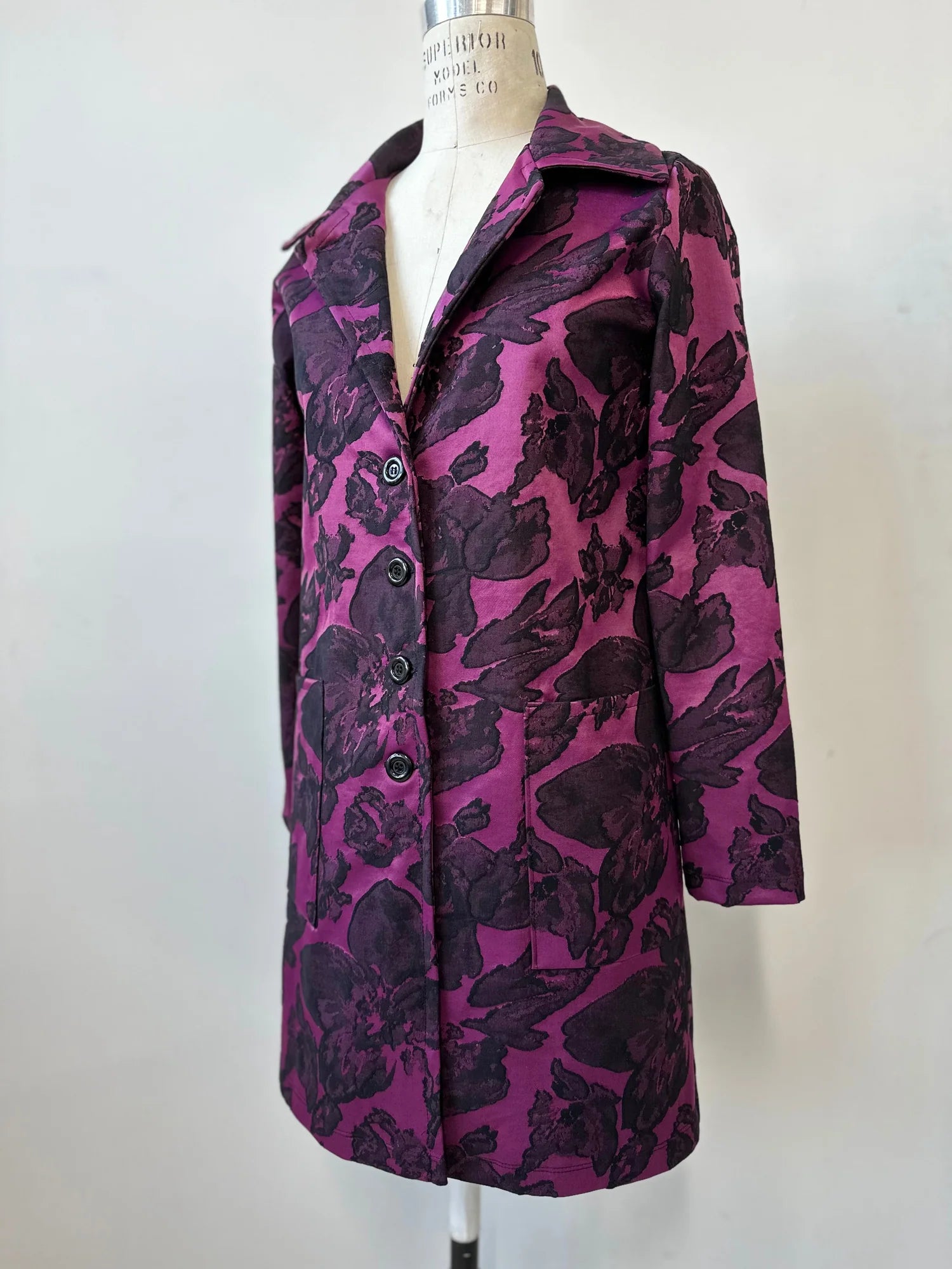 The Amarante Coat by Kollontai in Blackberry is shown on a mannequin against a white background. It's a satin coat with an intricate floral pattern, a classic collar, button front, straight cut that hits above the knee, and large pockets. 
