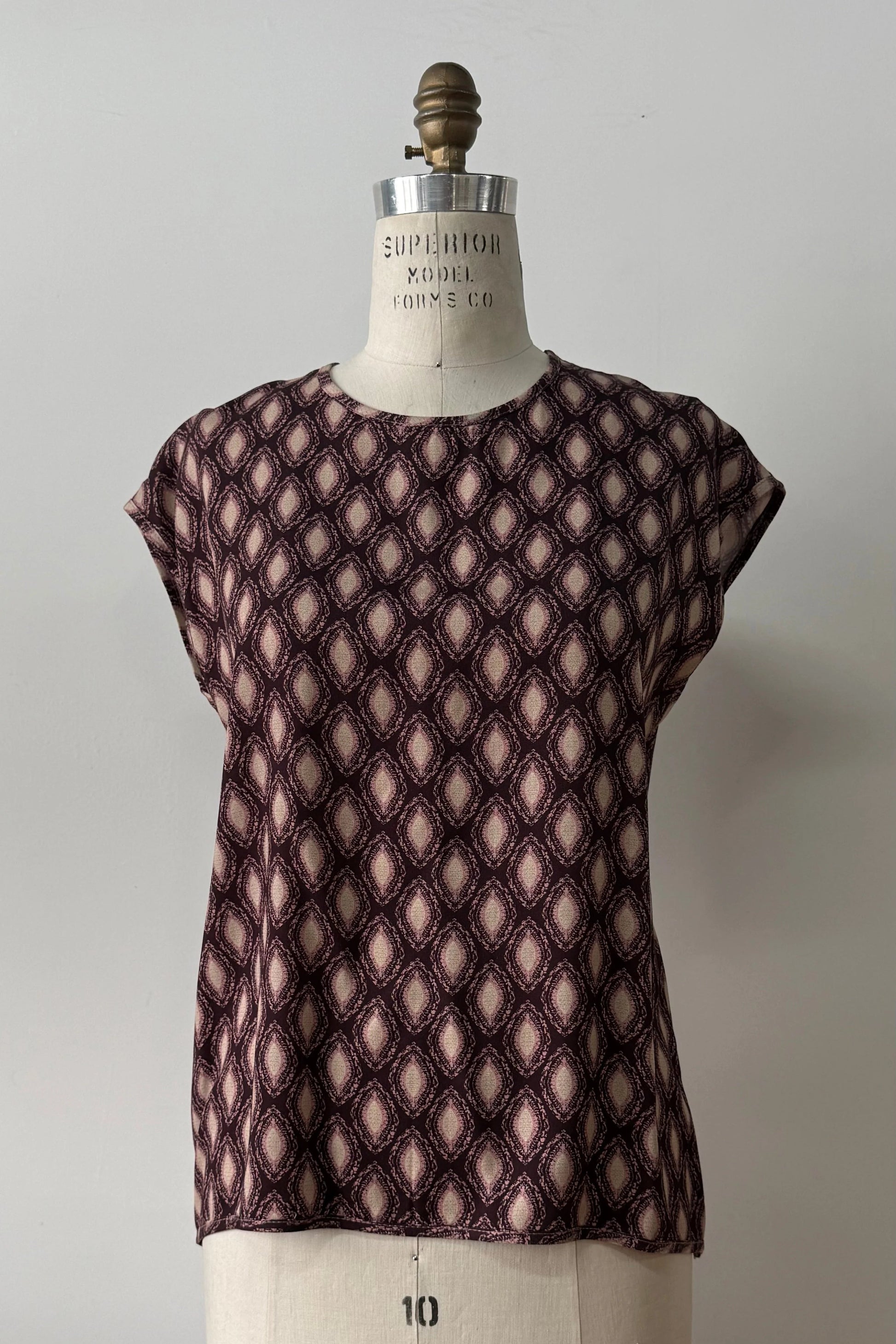 The Fanie Shirt by Kollontai, a relaxed fitting top with cape sleeves and a hi-low hem in a pink and plum geometric print, is shown on a mannequin against a white background. 
