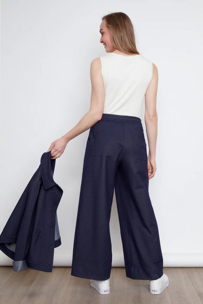 Back view of a woman wearing the Brooke Knit Trousers by Julei in Navy Knit, wide-leg pants with a pull-on waist, front slant pockets, and back patch pockets. She is wearing them with a white tank and holding a navy jacket against a white background.