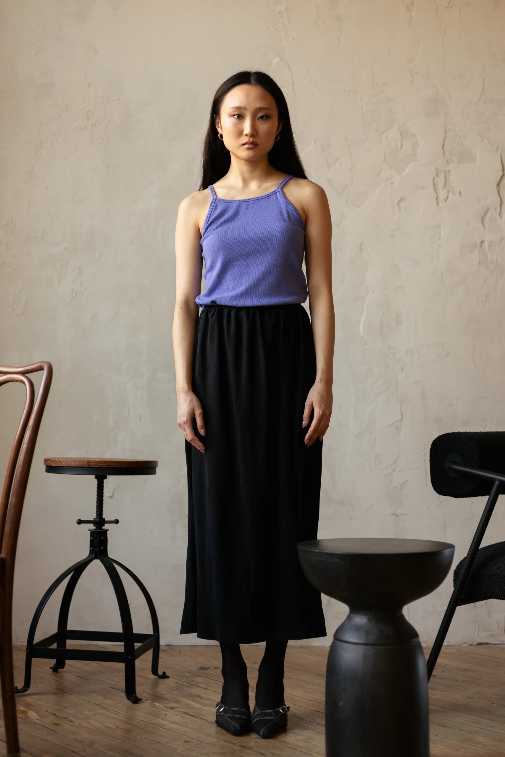 A woman wearing the Simone Skirt by Lights of All in Black, a sleek calf-length skirt with a flared hem and an elastic waist. She is wearing it with a purple top and standing in a room with furniture. 