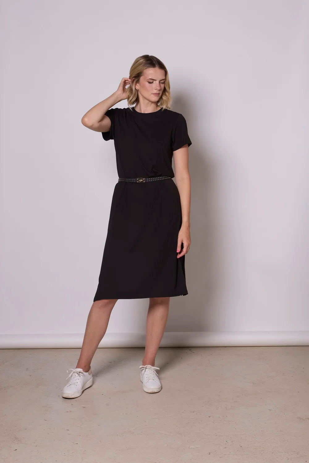 A woman wearing the Frances Midi  by Tangente in Black, a short sleeved loose fitting dress with side slits. She is wearing it with a black belt and standing against a plain background