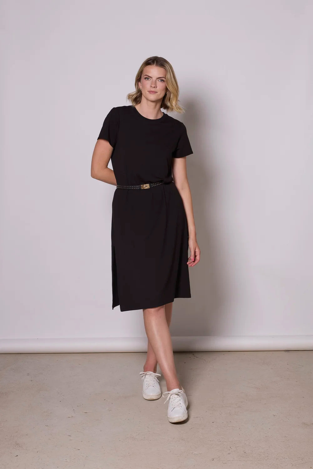 A woman wearing the Frances Midi  by Tangente in Black, a short sleeved loose fitting dress with side slits. She is wearing it with a black belt and standing against a plain background