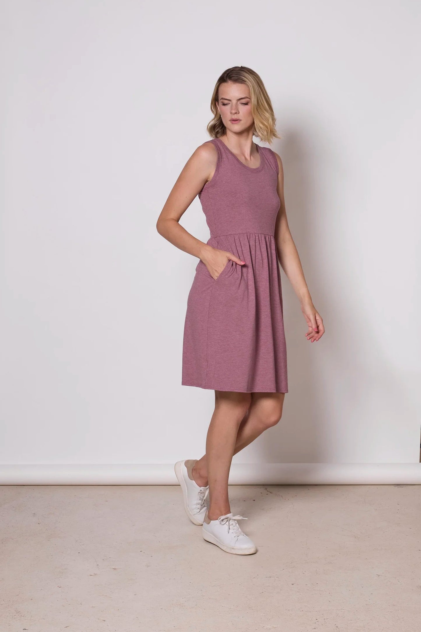 A woman wearing the Mai Dress by Tangente in Heather Rose, a  sleeveless dress with a scooped neck and gathered knee-length skirt. She is standing against a white background