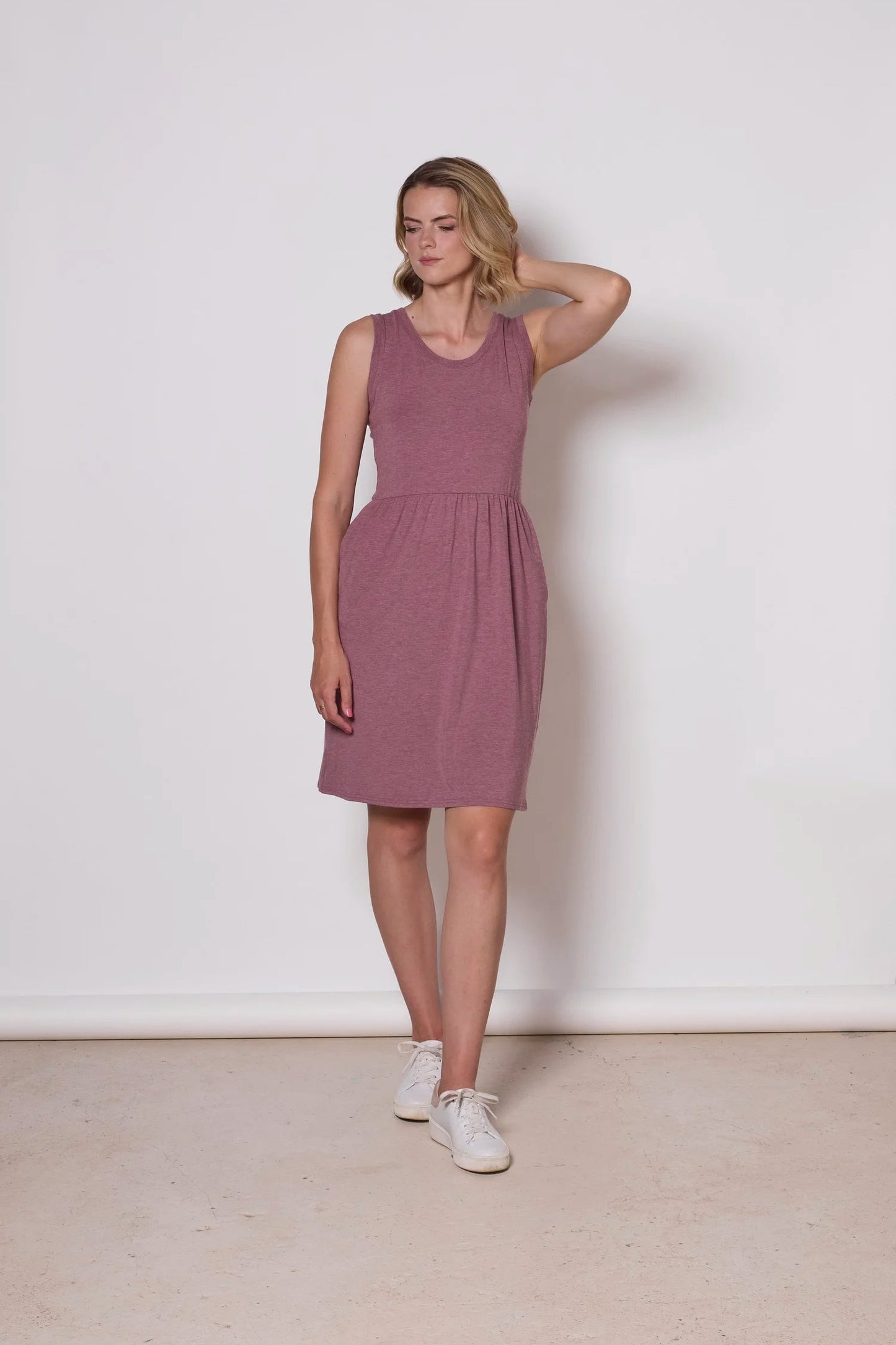 A woman wearing the Mai Dress by Tangente in Heather Rose, a  sleeveless dress with a scooped neck and gathered knee-length skirt. She is standing against a white background