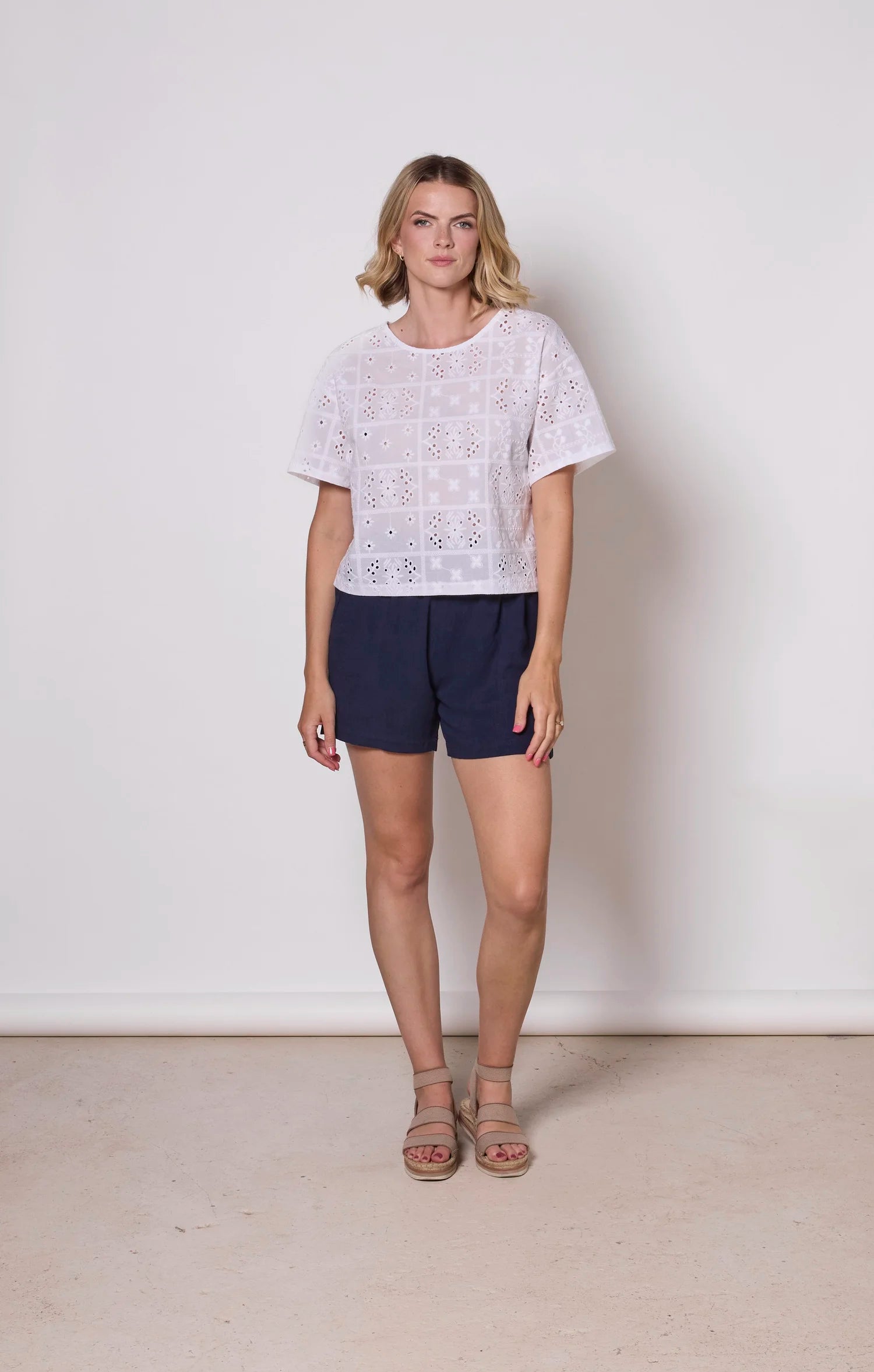 A woman wearing the Florence Top by Tangente in White, an embroidered eyelet top with short drop sleeves and a boxy fit. She is wearing it with navy shorts  against a plain background. 