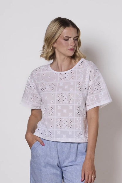 A woman wearing the Florence Top by Tangente in White, an embroidered eyelet top with short drop sleeves and a boxy fit. She is wearing it with light blue pants against a plain background. 