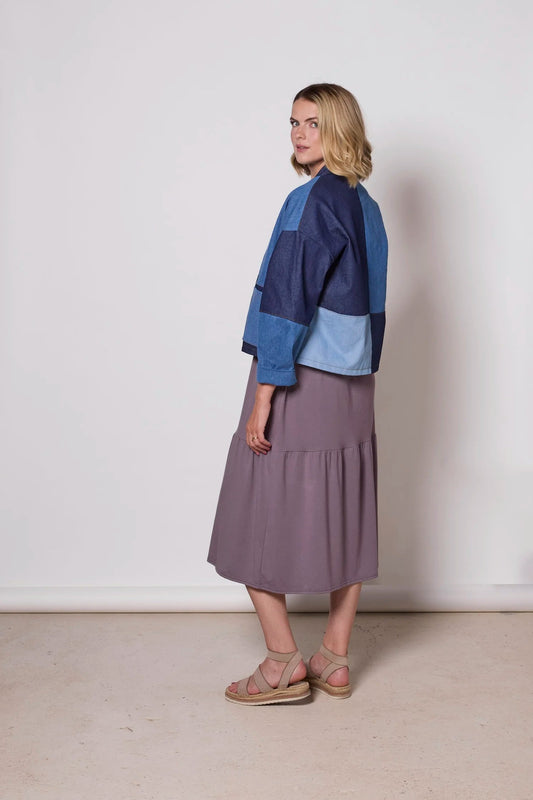 A woman wearing the Myrtle Denim Jacket by Tangente, a patchwork jacket with an open front, drop sleeves, and front patch pockets. She is wearing it over a purple dress against a white background