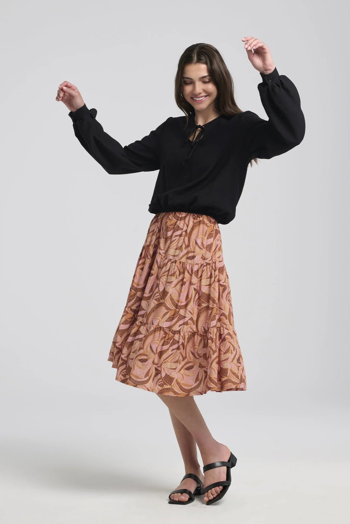 A woman wearing the Al-Uzza Skirt by Kollontai in Black, a peasant blouse with long puffed sleeves, a bloused hem, and a cut-out and tie detail at the front neckline. She is wearing it with the Al-Uzza Skirt in Cinnamon print and standing in front of a white background. 