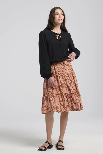 A woman wearing the Al-Uzza Skirt by Kollontai in Black, a peasant blouse with long puffed sleeves, a bloused hem, and a cut-out and tie detail at the front neckline. She is wearing it with the Al-Uzza Skirt in Cinnamon print and standing in front of a white background. 