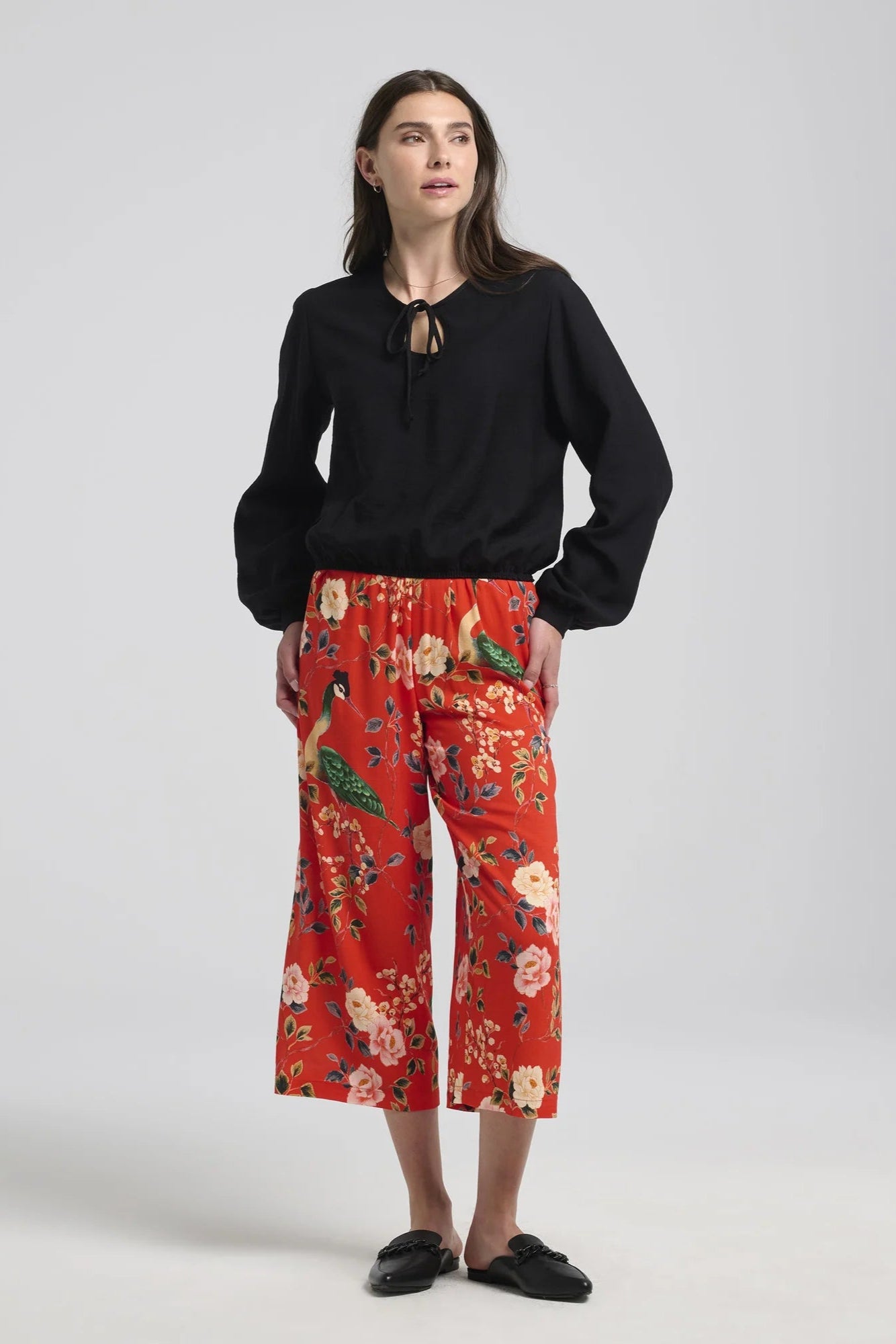 A woman wearing the Al-Uzza Skirt by Kollontai in Black, a peasant blouse with long puffed sleeves, a bloused hem, and a cut-out and tie detail at the front neckline. She is wearing it with the Paraiso Pants and standing in front of a white background. 
