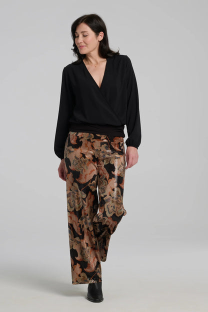A woman wearing the Carina Pants by Kollontai, featuring floral velvet in autumn tones with clear sequins sewn right into the fabric. They have full-length straight legs and a wide pull-on waistband. She's wearing them with a black top and standing in front of a grey background. 