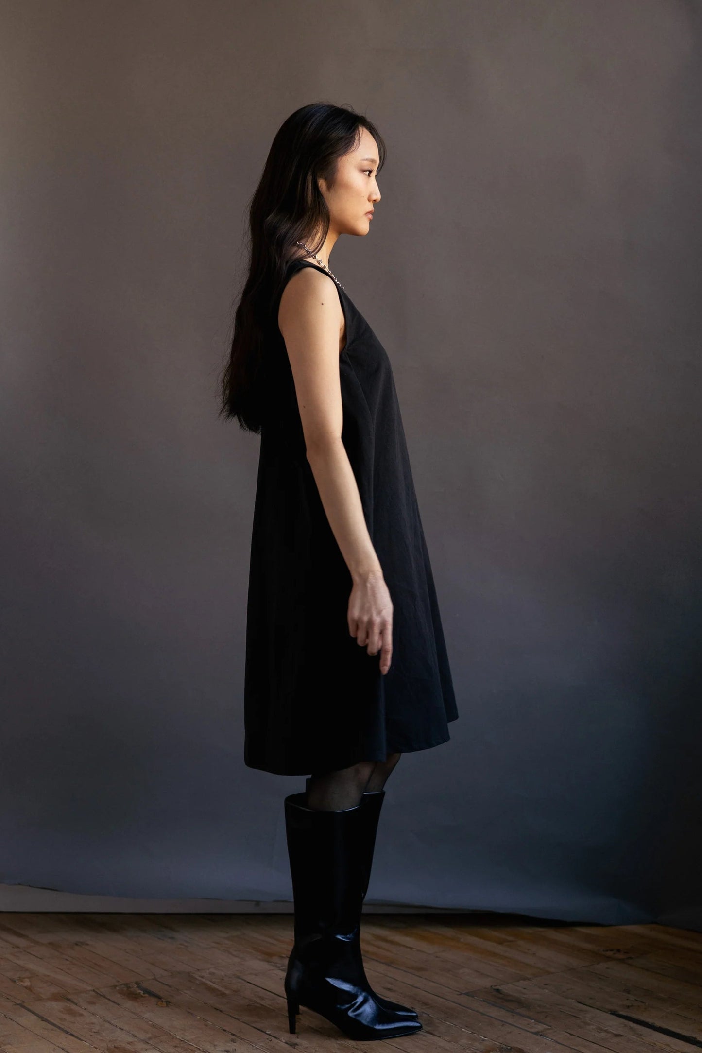 A woman wearing the Caro Dress by Lights of All in Black, a sleeveless knee-length trapeze dress with a round neck at the front and a V at the back. She is wearing it with knee-high black boots standing in front of a dark backdrop.
