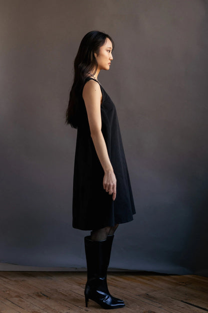 A woman wearing the Caro Dress by Lights of All in Black, a sleeveless knee-length trapeze dress with a round neck at the front and a V at the back. She is wearing it with knee-high black boots standing in front of a dark backdrop.