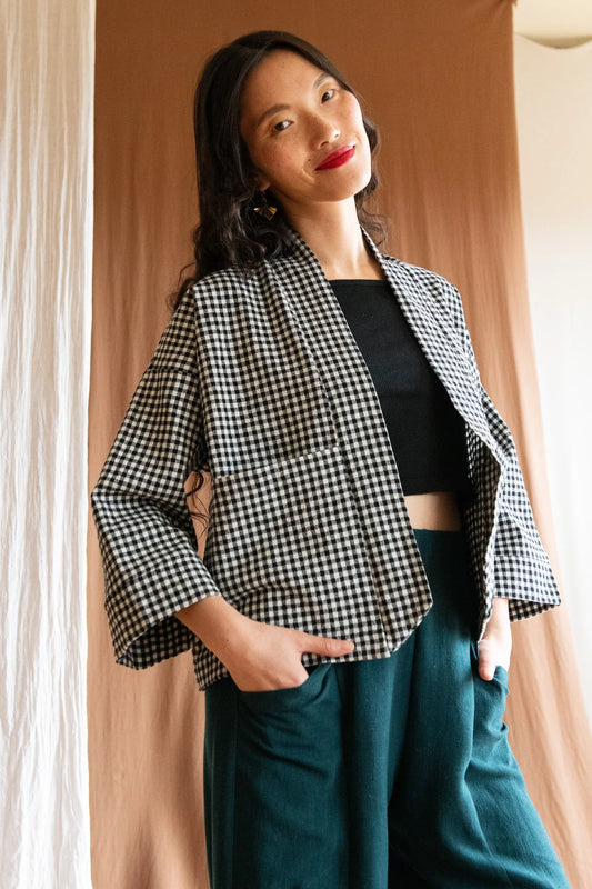 3/4 shot of a woman wearing the Pisa Jacket by Kazak in Black and White Gingham, a short kimono-style jacket with an open front, angled hem, wide 3/4 sleeves and large pockets. She is wearing it with the Padua Pants in Forest and standing in front of a brown curtain. 