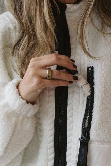 Close up of a person wearing a white fleece jacket tucking Picot's Honey Tobacco fragrance rollerball into the chest pocket. 