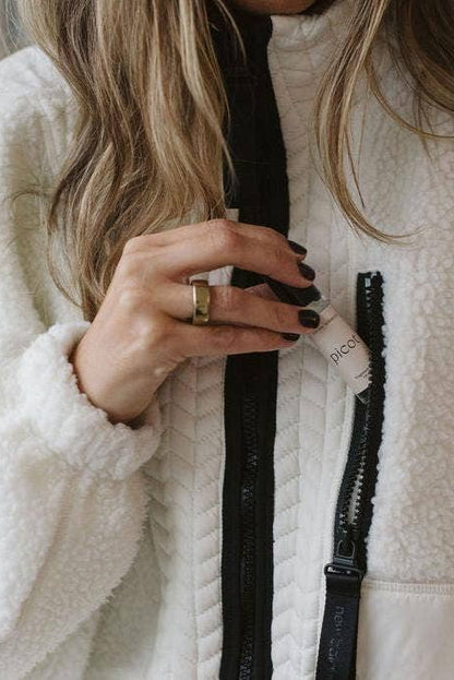 Close up of a person wearing a white fleece jacket tucking Picot's Honey Tobacco fragrance rollerball into the chest pocket. 